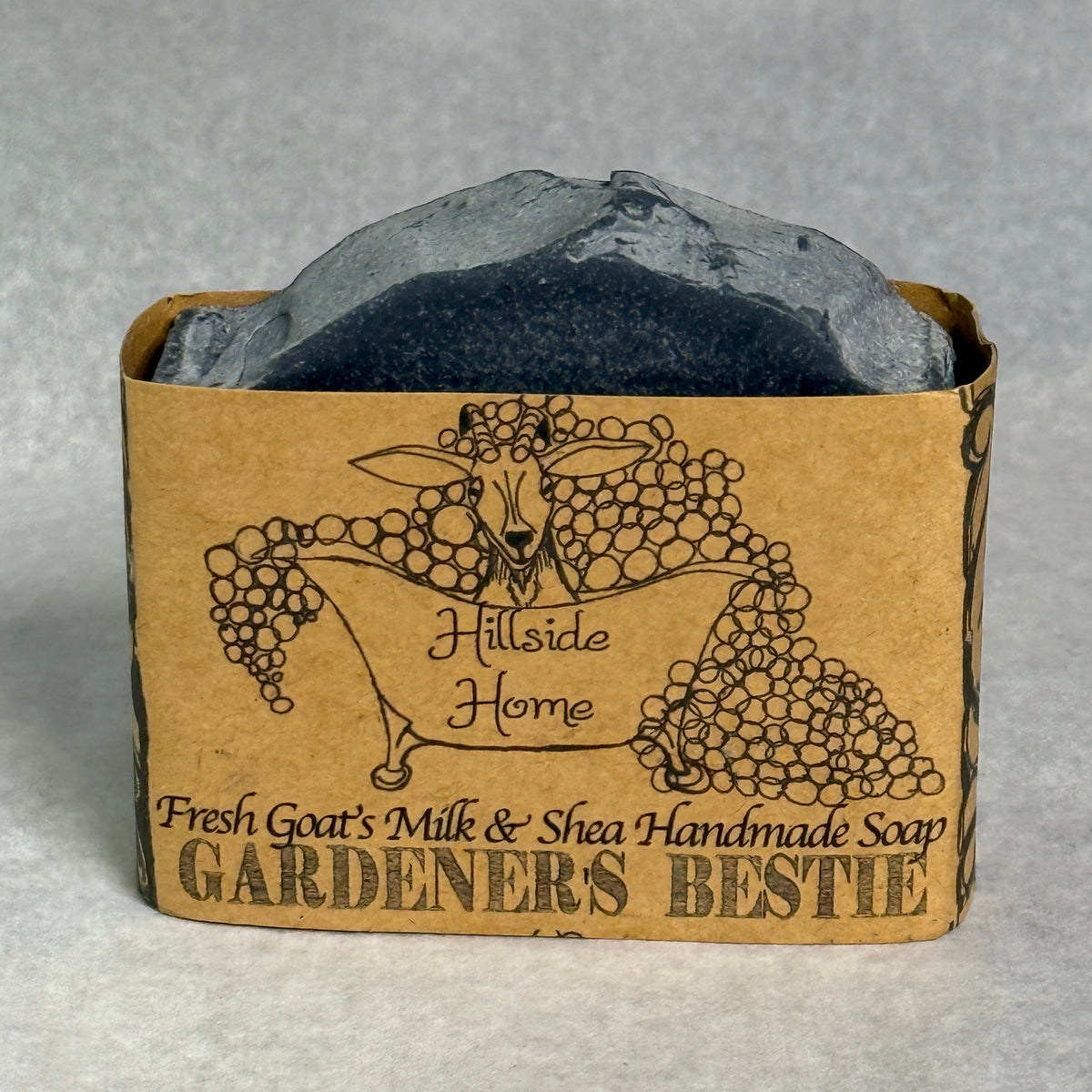 A rectangular bar of handmade soap in a brown kraft paper sleeve labeled 'GARDENERS BESTIE' by Hillside Home, with a dark charcoal soap bar visible at the top against a light gray background.