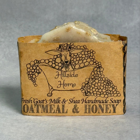 A handmade soap bar wrapped in a kraft paper sleeve labeled 'Hillside Home Fresh Goat's Milk & Shea Handmade Soap Oatmeal & Honey,' photographed on a neutral gray background.