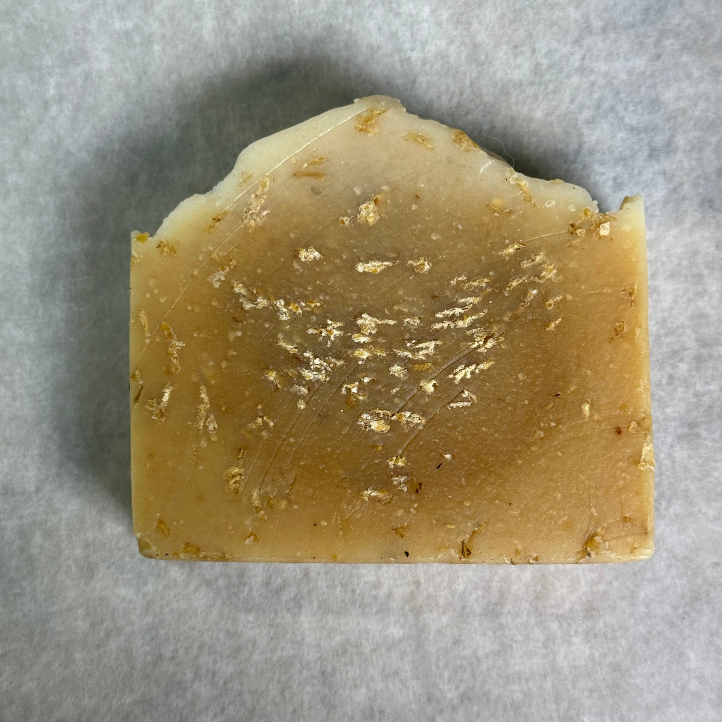 A rectangular bar of handmade soap with a rustic, irregular top edge, featuring a warm tan and light brown color with scattered small gold flakes, set against a neutral gray background.